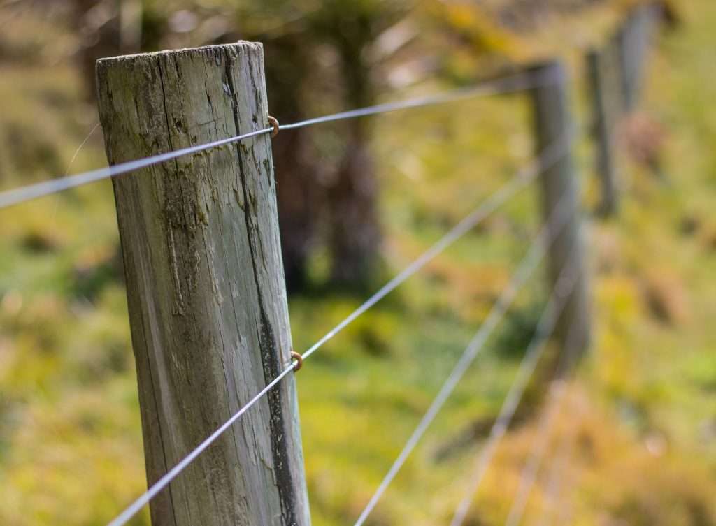 Close-up of a wooden fencing post with four metal wires running horizontally through it, set in a grassy farm area with a blurred background.