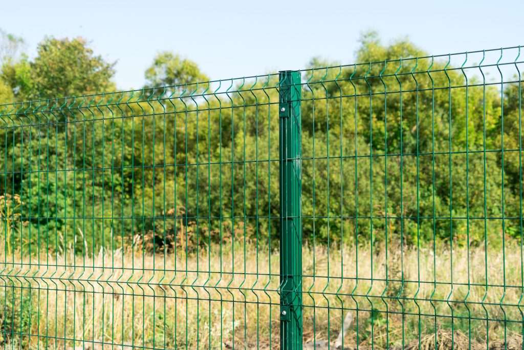 A green metal wire fence with a vertical support post stands in front of a grassy farm field and trees under a clear sky, perfect for fencing needs.